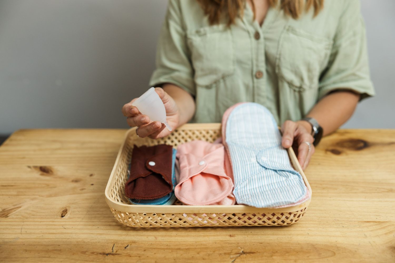 A close-up shot of a person holding a silicone cup above a shallow woven basket containing an assortment of colorful, reusable cloth pads on a wooden table.